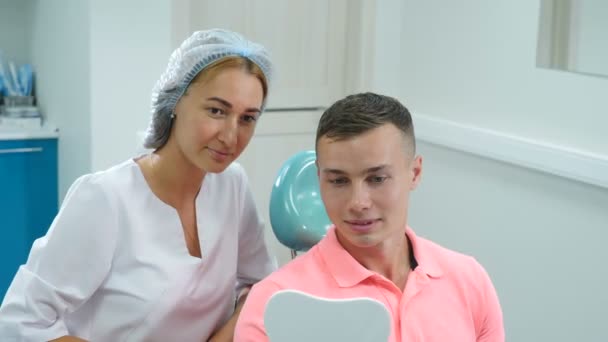 Позитивный красивый мужчина пациент стоматологической клиники наслаждается удивительной улыбкой после лечения. Женщина-врач и молодой человек улыбаются, глядя друг на друга. Гигиена полости рта в современной стоматологии. Человек смотрит в зеркало — стоковое видео