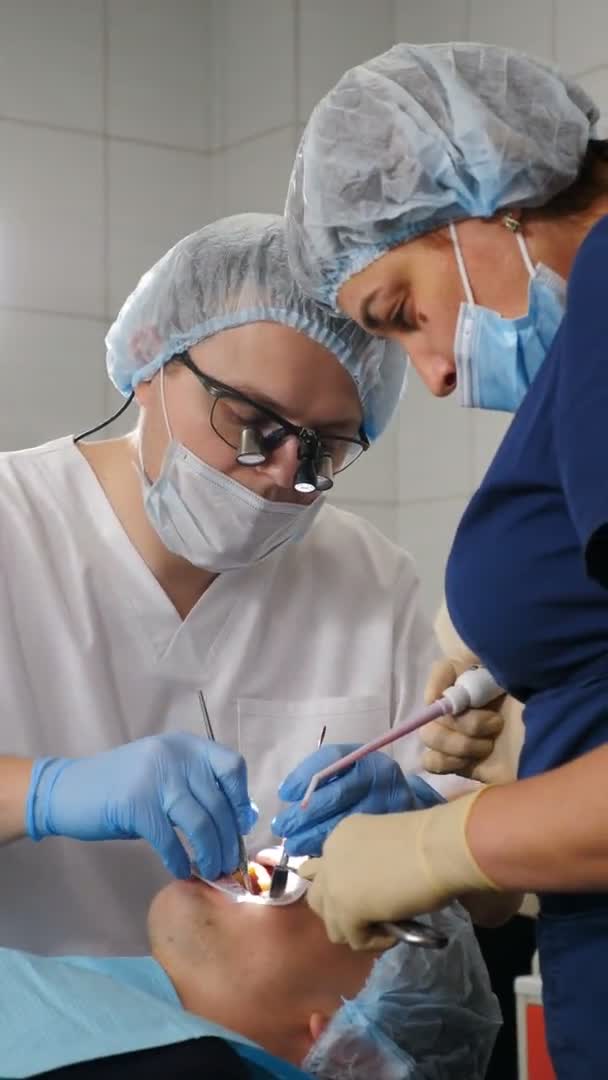 Modern tandläkarmottagning. Vertikal video. Professionell tandläkare kirurg behandla rotkanaler, extrahera tand eller installera tandimplantat. Läkare och assistent bära skyddsdräkt och handskar arbetar på — Stockvideo