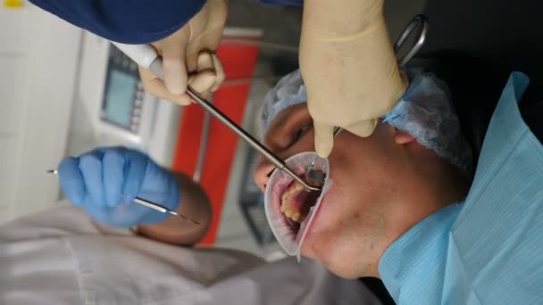 Modern tandkirurgi på kliniken. Professionell tandläkare kirurg behandla rotkanaler, extrahera tand eller installera tandimplantat. Vertikala bilder. Läkare och assistent slitage skyddsdräkt arbetar — Stockvideo