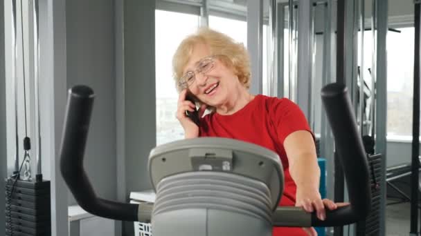 携帯電話で話す運動自転車の練習に従事する笑顔の高齢女性。リハビリテーションセンタージム怪我や骨折からの回復。シミュレータ上で運動を行うシニア女性。四キロ — ストック動画