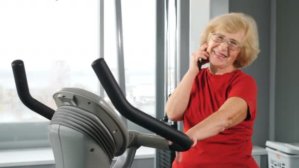 Uśmiechnięta starsza kobieta zaangażowana w trening kardiologiczny na rowerze treningowym rozmawiająca przez telefon. Siłownia ośrodka rehabilitacyjnego, rekonwalescencja po urazach i złamaniach. Starsza kobieta wykonuje ćwiczenia na symulatorze. 4 k — Wideo stockowe