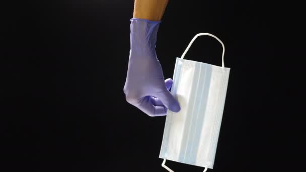 Hand in Hand mit blauem Schutzhandschuh hält medizinische Maske gegen Viren und Bakterien auf schwarzem Hintergrund, Schutz gegen neue Welle des Coronavirus, COVID-19. Medizin und Gesundheitskonzept. Vertikales Video — Stockvideo