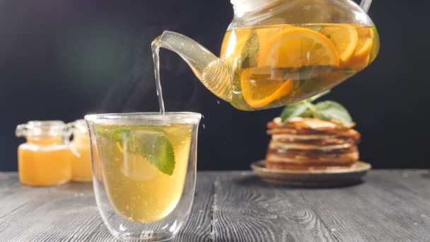 Verser le thé aux fruits avec de l'orange, du citron et du citron vert de la théière transparente dans un verre avec une menthe. Set de thé, théière en verre et tasses. Mélange de thé avec du miel et des crêpes en arrière-plan. Vidéo au ralenti — Video