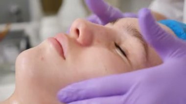 Güzellik uzmanı, kadın müşterinin yüzüne asit soyma çözeltisi sürüyor eldivenli ellerle ovuyor. Cilt bakımı ve sorunlu cilt için prosedürler. Genç bir kadın güzellik kliniğinde yüz bakımı yaptırıyor. 4 bin.