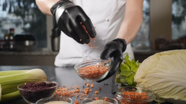 Chef en gants noirs saupoudrer une poignée de carottes tranchées séchées. Épices et ingrédients alimentaires. Au ralenti. Mélange d'espèces séchées colorées et de légumes sont en baisse. Carotte nettoyée, lavée, séchée — Video