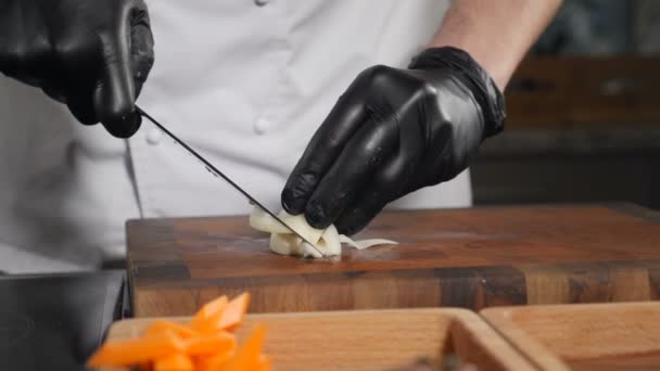Готуємо домашню їжу, нарізаємо овочі. Шеф-кухар в чорних рукавичках нарізає цибулю ножем. Повільний рух. Процес приготування їжі. Шеф-кухар подрібнює цибулю на маленькі скибочки на дерев'яній обробній дошці. Повний HD — стокове відео