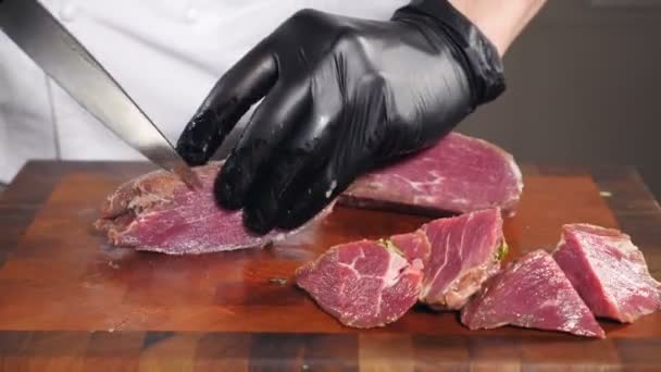 黒手袋のシェフは、木製のまな板にナイフで生肉をカットします。料理の前にテーブルの上の牛肉や仔牛の肉。手袋の肉屋は肉を部分に切る。鋭いナイフで生肉を切る。四キロ — ストック動画