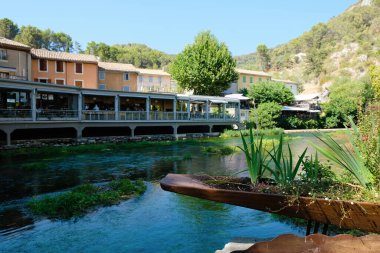 Fontaine de Vaucluse, Provence, Fransa. 09 Ağustos 2022. Sorgue Nehri 'ndeki dekoratif tekne çiçek yatağı ve restoran arka planı. Ünlü bir yer..