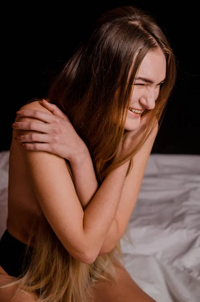 Piękna młoda kobieta z blond włosami w czarnej bieliźnie siedzi na łóżku, uśmiechnięta, estetyczne pozy, czarne tło — Zdjęcie stockowe