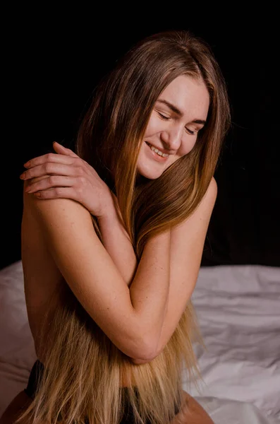Piękna młoda kobieta z blond włosami w czarnej bieliźnie siedzi na łóżku, uśmiechnięta, estetyczne pozy, czarne tło — Zdjęcie stockowe