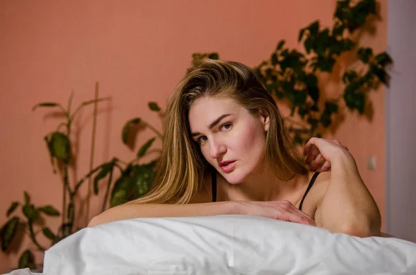 Όμορφη νεαρή γυναίκα με ξανθά μαλλιά ακουμπισμένα στο κρεβάτι σε αισθητικές πόζες, και υπάρχουν πράσινα φυτά σε καφέ φόντο — Φωτογραφία Αρχείου