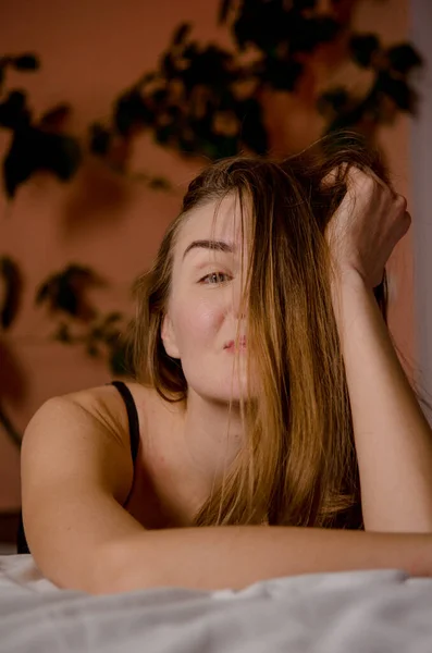 Gyönyörű fiatal nő szőke haj támaszkodik az ágyra esztétikai pózok, és vannak zöld növények barna háttér — Stock Fotó