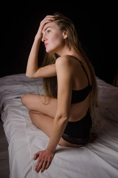 Krásná mladá žena s blond vlasy v černém spodním prádle sedí na posteli v estetické pózy, na černém pozadí — Stock fotografie