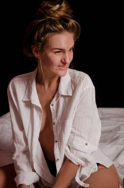 Gyönyörű fiatal nő szőke hajjal, fehér ingben, meztelen testen, fehér lepedőn ül esztétikai pózokban. — Stock Fotó