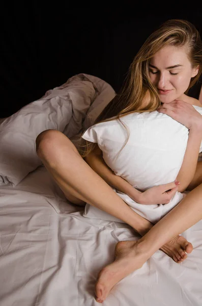 Beautiful thin young woman with long blonde hair in a white bed sits hugging a pillow and laughs