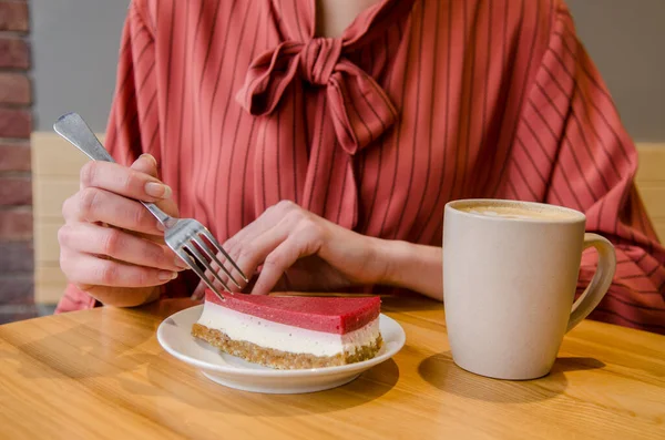 Young woman in red dress sits at table in cafe, she eats red cake on saucer with a fork, cup with drink is nearby