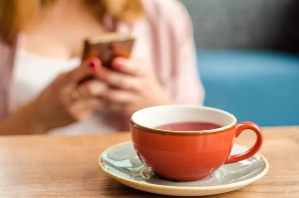 Red cup of red tea, a gray teapot stand on a table in a cafe. In the background, a young woman is working on her smartphone