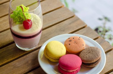 Büyük şeffaf bardakta vişneli tatlı, beyaz çay tabağında farklı türde makarna pastası, açık kahvenin terasında duruyor, arka plandaki ahşap masada ve güneşli bir günde