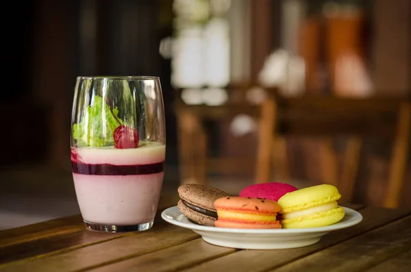 Large transparent glass with cherry dessert, different types of macaron cake on a white saucer standing on the outdoor terrace of cafe, on a wooden table against background of the windows of cafe