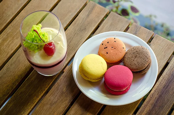Large transparent glass with cherry dessert, different types of macaron cake on white saucer standing on the outdoor terrace of cafe, on a wooden table against the background greenery and a sunny day