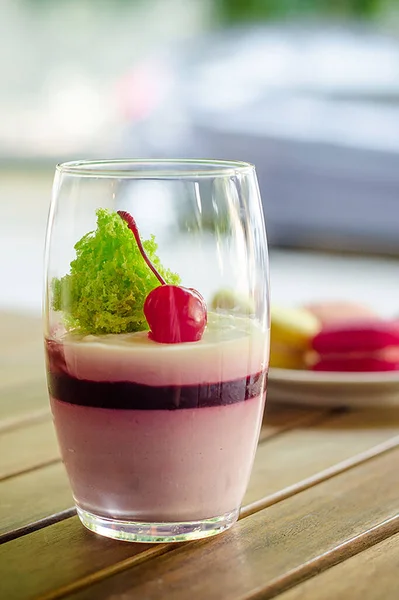 Large transparent glass with cherry dessert, different types of macaron cake on white saucer standing on the outdoor terrace of cafe, on a wooden table against the background greenery and a sunny day