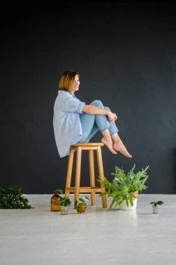 Mavi gömlekli ve kot pantolonlu genç Avrupalı bir kadın, çıplak ayaklı, ahşap bir taburede oturuyor, siyah arka planda yeşil bitkilerle çevrili.