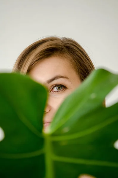 年轻的欧洲女人 金发碧眼 穿过迷蒙的月季植物的大叶 在一个白光的房间背景上 — 图库照片