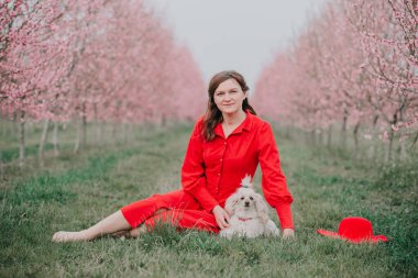 Bir kadın köpeğiyle çimlerin üzerinde oturuyor. Bahar bahçesi çiçek açıyor..