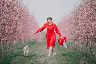 Bir kadın ve köpeği bahçede yürüyorlar. Ağaçlar çiçek açıyor. İlkbahar.