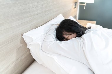 Beyaz kollu, uzun kollu Asyalı kadın pijamaları yüzükoyun yatar, yatakta, denenmiş ve yorgun ruh hali ve tonu ile yüzüstü yatar..