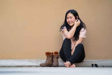 Rahat, güzel Asyalı kadın, açık kahverengi arka planıyla çatıda oturuyor ve kameraya gülümsüyor..