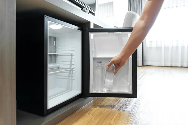 Man Hand opened mini small refrigerator and reached drinking plastic water bottle under the wood counter in the hotel resort bedroom.