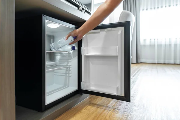 Man Hand opened mini small refrigerator and reached drinking plastic water bottle under the wood counter in the hotel resort bedroom.