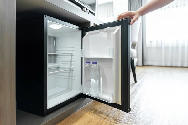 Man Hand opened mini small refrigerator and reached drinking plastic water bottle under the wood counter in the hotel resort bedroom.