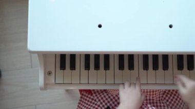A girl in long sleeved red and white plaid dress is playing a little white piano with her parent beside.