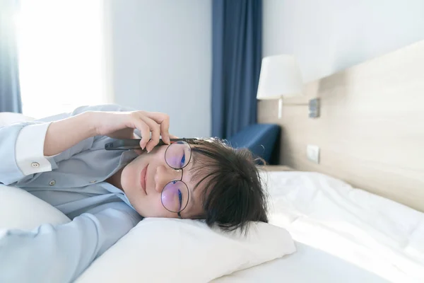 Asian young glasses woman talk to the mobile phone while lie on her side on the bed in the morning.