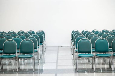 Geniş bir salonda bir sürü boş yeşil deri sandalye, ışıklar açık. Zemin nesneleri yansıtır. Boşluk hissi verir.  