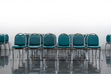 Geniş bir salonda bir sürü boş yeşil deri sandalye, ışıklar açık. Zemin nesneleri yansıtır. Boşluk hissi verir.  