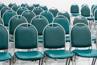 Geniş bir salonda bir sürü boş yeşil deri sandalye, ışıklar açık. Zemin nesneleri yansıtır. Boşluk hissi verir.  