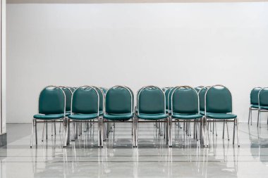 Geniş bir salonda bir sürü boş yeşil deri sandalye, ışıklar açık. Zemin nesneleri yansıtır. Boşluk hissi verir.  