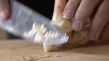 Galangal, ahşap bir tahtanın üzerinde kesilip küçük parçalara bölünüyor ve profesyonel bir şef tarafından mutfak bıçağıyla dilimleniyor..