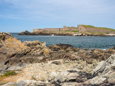 Fort Doyle 'dan Fort Grosnez' e kayalık körfezin karşısındaki mavi gökyüzü ve beyaz bulutların altında Alderney, Channel Adaları 'na bakın.