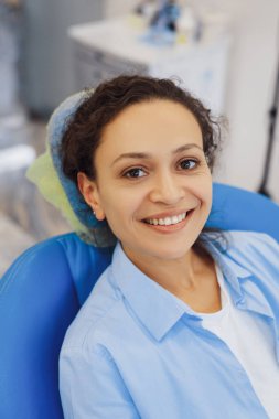 Pozitif kadın hasta dişçiyi ziyaret ediyor ve modern klinikte kameraya gülümsüyor.