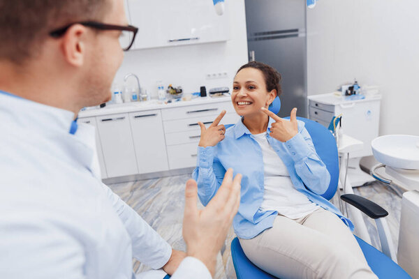 Веселая женщина указывает пальцем на здоровые зубы. Мужчина-дантист беседует с клиенткой на стоматологическом стуле современной клиники