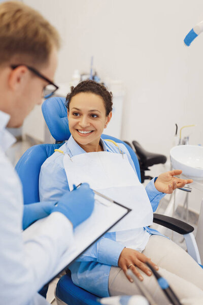 Smiling woman talking with dentist in dentistry medical room. Male dental doctor making notes on clipboard