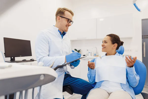 Happy dental doctor writing diagnosis on clipboard while smiling woman sitting in dental hair