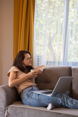 Genç bir kadın evden çalışırken bir fincan çay molası verir. Bir kız elinde dizüstü bilgisayarla kanepede oturuyor.