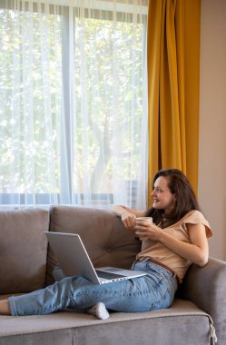 Bir kadın evde çalışırken bir fincan çaya ara verir. Bir kız kanepede dizüstü bilgisayarla oturuyor ve gülümsüyor.