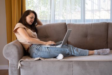 Evden çalışan genç bir kadın. Bir kız elinde dizüstü bilgisayarla kanepede yatıyor.