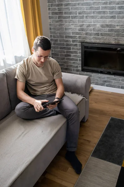 A young man is sitting on a sofa and playing on a tablet. Guy typing something — Stock Photo, Image
